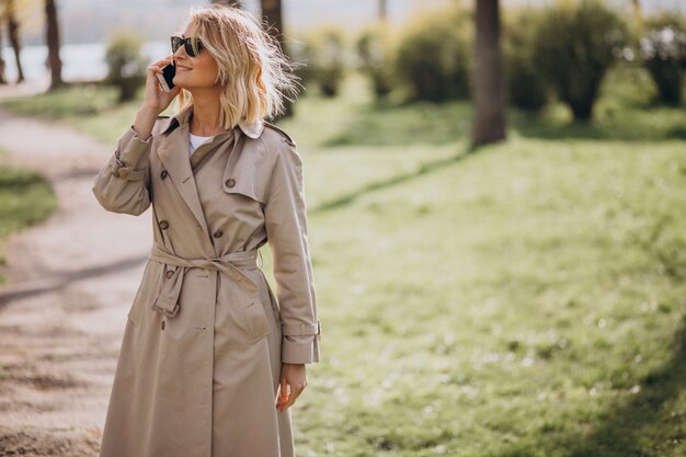 Белокурая женщина в пальто снаружи в парке используя телефон