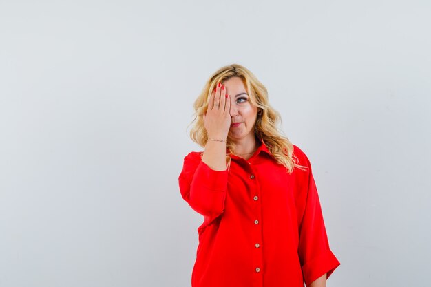 Blonde woman covering eye with one hand in red blouse and looking happy.