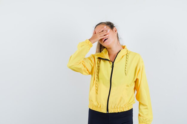 Blonde woman covering eyes with hand in yellow bomber jacket and black pants and looking harried