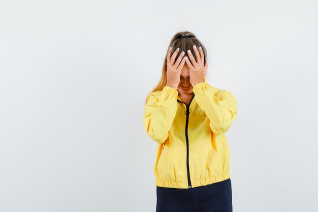 Blonde woman covering head with hands in yellow bomber jacket and black pants and looking harried