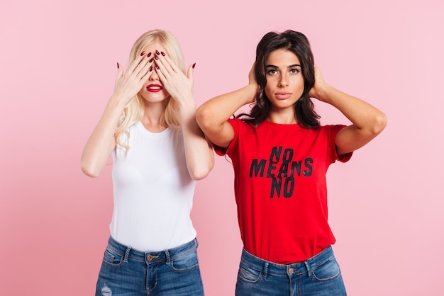 Free photo blonde woman covering her eyes and brunette woman covering her ears over pink