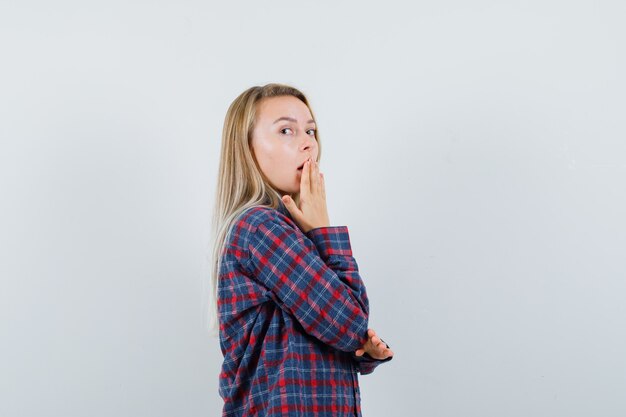 Blonde woman covering mouth with hand in checked shirt and looking surprised , front view.