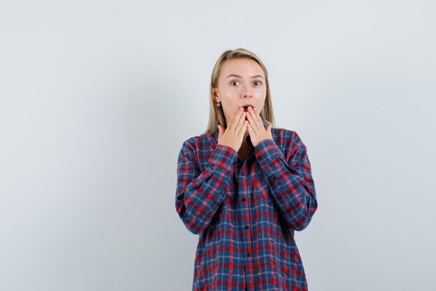 Blonde woman covering mouth with hands in checked shirt and looking surprised. front view.