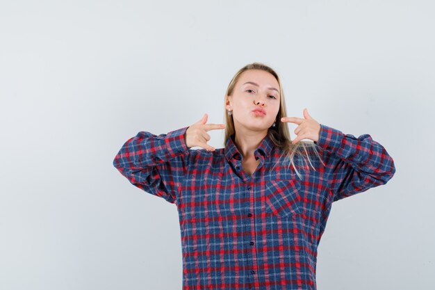 Blonde woman curving lips and pointing to it with index fingers in checked shirt and looking confident, front view.