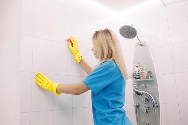 Блондинка, сотрудница клининговой компании, в желтых защитных резиновых перчатках со специальной чистящей губкой, очищающая поверхность плитки на стене в белой ванной
