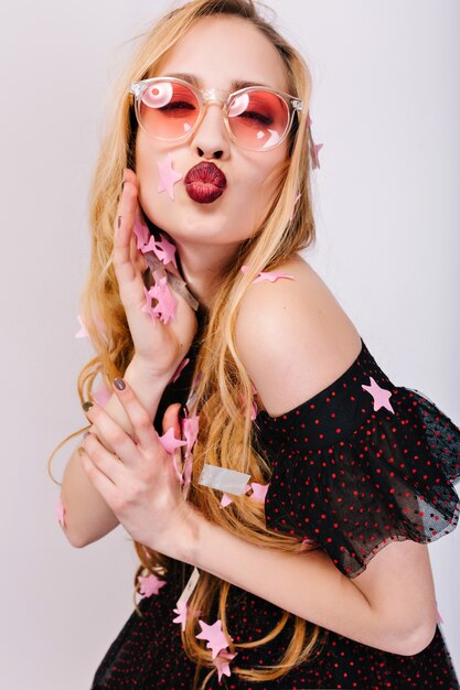 блондинка женщина, дающая поцелуй, весело на вечеринке, праздничное мероприятие, покрытая конфетти. В розовых очках, в красивом черном платье, длинные волосы.