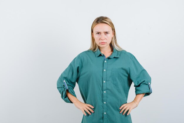 Blonde woman in green shirt holding hands on waist and looking serious