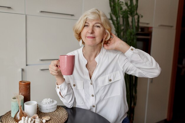 Blonde woman holding cup of tea and sitting at modern kitchen