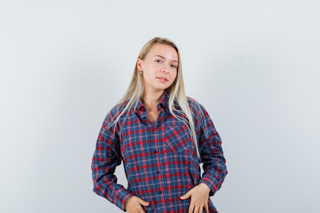 Blonde woman holding hands on belly, posing at camera in checked shirt and looking charming . front view.