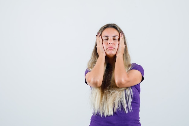 blonde woman holding hands to head in violet t-shirt and looking exhausted, front view.