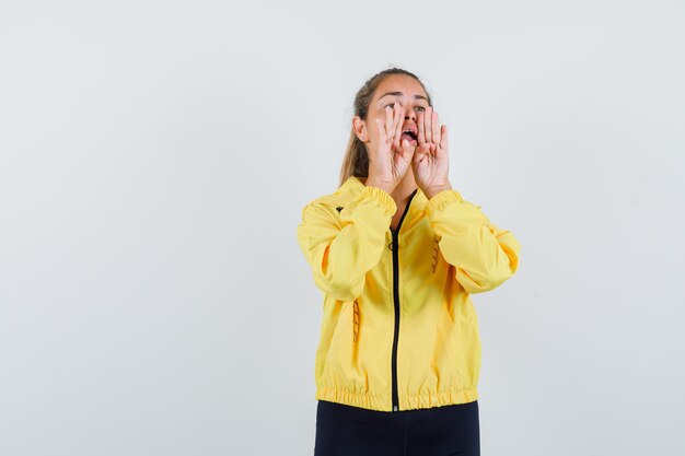 Blonde woman holding hands near mouth as calling someone in yellow bomber jacket and black pants and looking happy