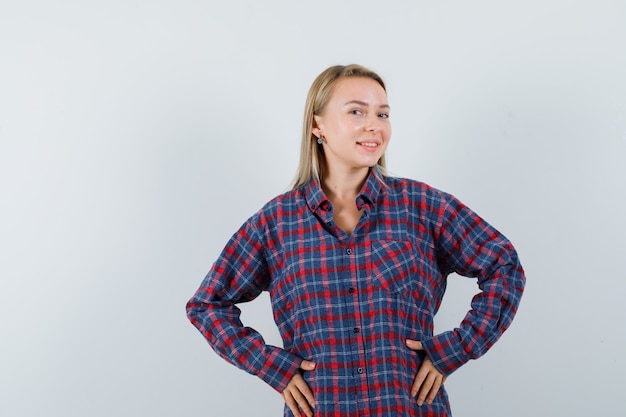 Free photo blonde woman holding hands over waist in checked shirt and looking optimistic , front view.