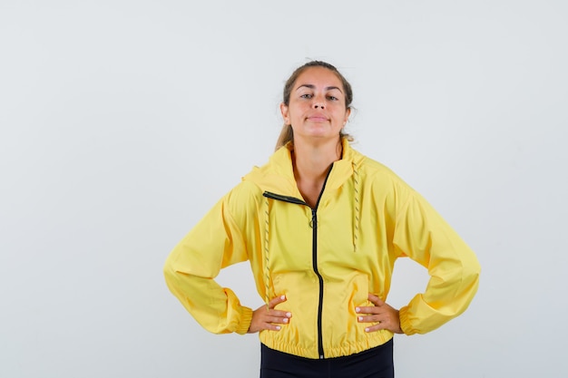 Free photo blonde woman holding hands on waist in yellow bomber jacket and black pants and looking happy