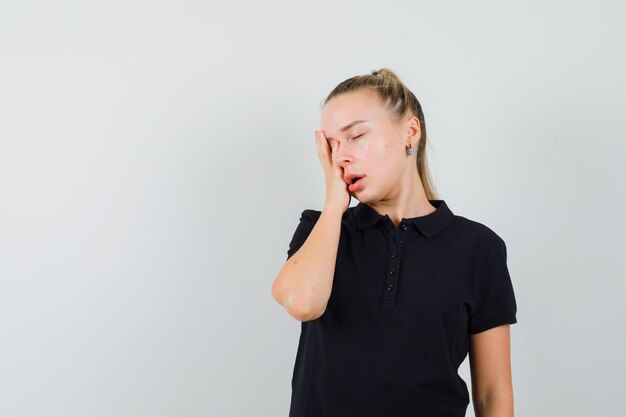 Blonde woman holding her hand on her head in black t-shirt and looking exhausted