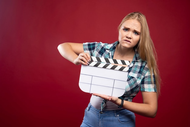 Blonde woman holding a movie production clapper board and looks confused and tired.