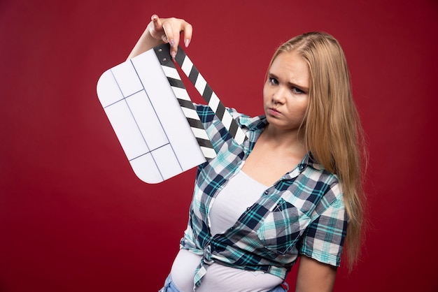 Free photo blonde woman holding a movie production clapper board and looks confused and tired.