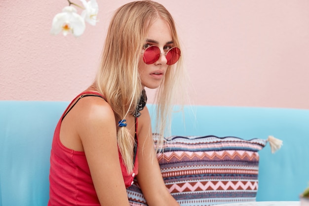 Бесплатное фото Блондинка в модных солнцезащитных очках, носит модную одежду и красные солнцезащитные очки, сидит у розовой стены на удобном диване.
