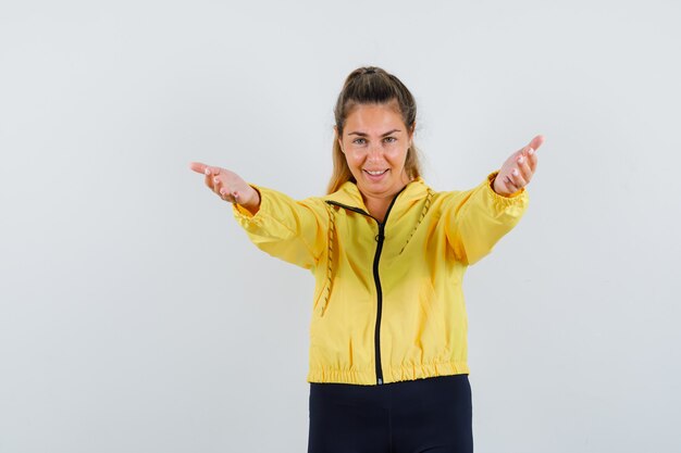 Blonde woman inviting to come in yellow bomber jacket and black pants and looking happy