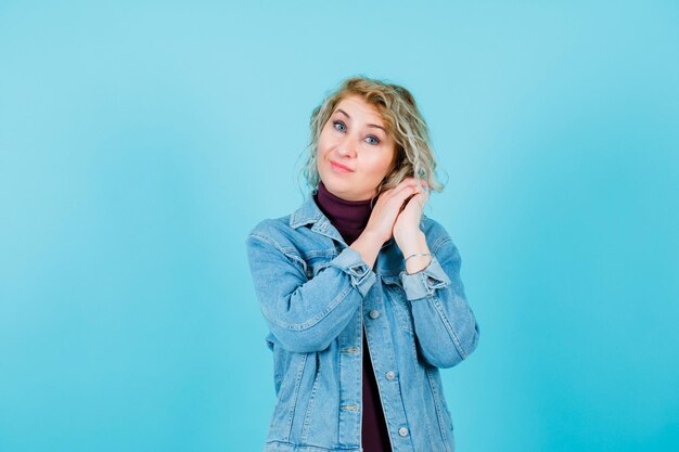 Blonde woman is holding hands together near ear on blue background