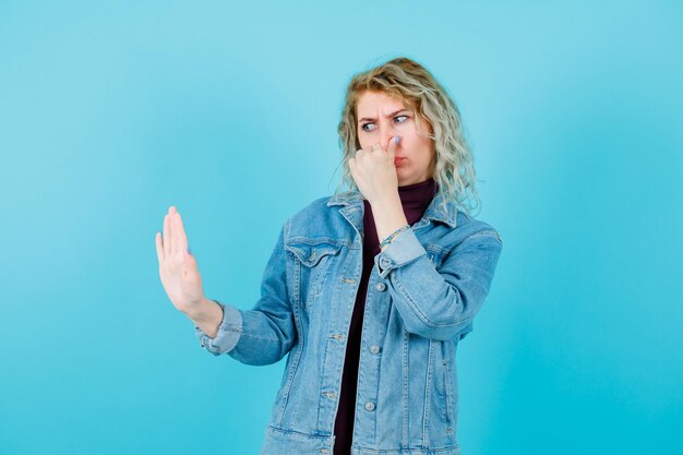 Blonde woman is holding nose hand showing stop gesture on blue background