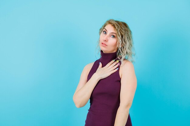 Blonde woman is looking at camera by holding hand on chest on blue background