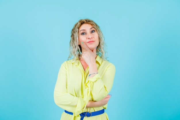 Blonde woman is looking at camera by holding hand on chin on blue background