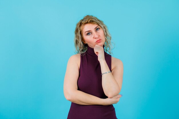 Blonde woman is looking at camera by holding hand on chin on blue background