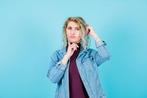 Blonde woman is looking at camera by holding one hand on ead and other hand on cheek on blue background