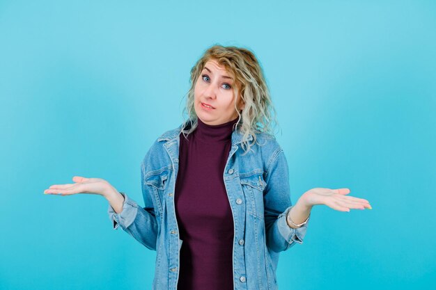 Blonde woman is looking at camera by opening wide her hands on blue background