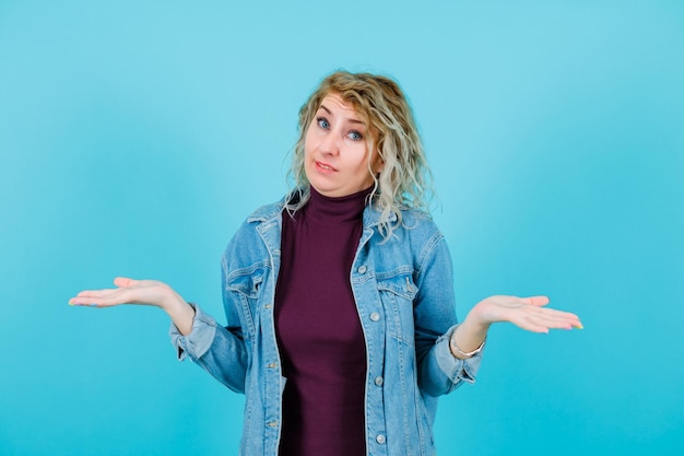 Blonde woman is looking at camera by opening wide her hands on blue background