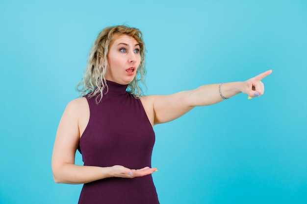 Blonde woman is pointing away by looking away on blue background