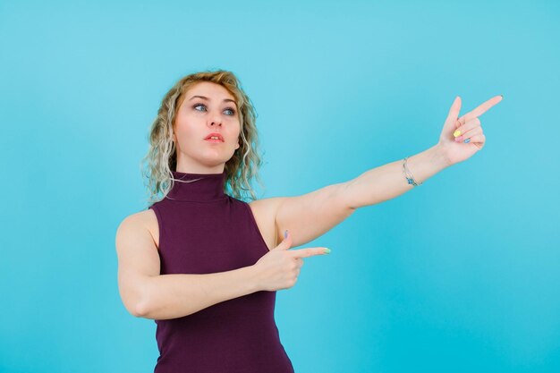 Blonde woman is pointing right up with forefingers on blue background