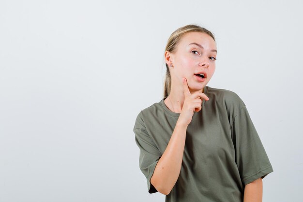 The blonde woman is putting her forefinger on cheek on white background