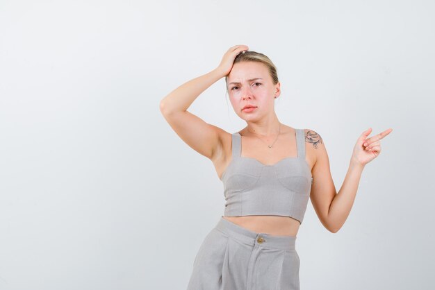 The blonde woman is putting her hand on head and pointing to right with other on white background