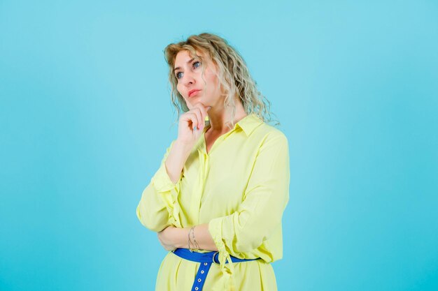 Blonde woman is thinking by putting hand on chin on blue background