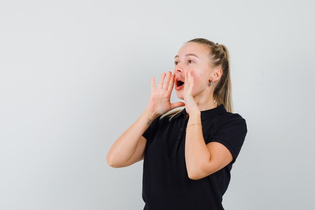 Blonde woman keeping her mouth wide open,trying to shout in black t-shirt and looking happy