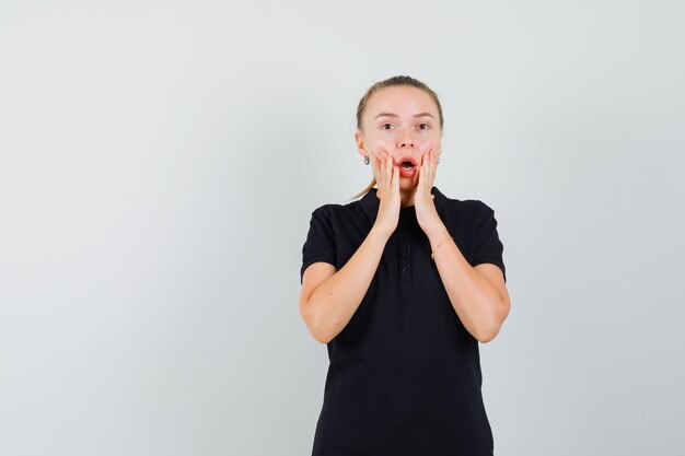 Blonde woman keeping mouth wide open, putting hands on cheeks in black t-shirt and looking shocked