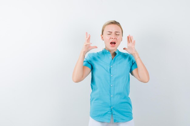 Blonde woman keeping raised hands near head in blue blouse and looking wistful