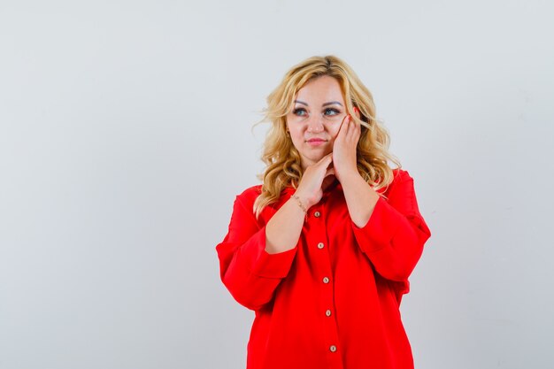 Blonde woman leaning cheek on hand, posing at camera in red blouse and looking pretty