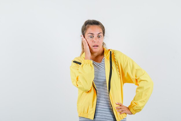 Blonde woman leaning cheek on palm while holding hand on waist in yellow bomber jacket and striped shirt and looking surprised