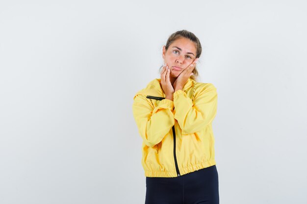 Blonde woman leaning cheek on palm while putting another hand on cheek in yellow bomber jacket and black pants and looking serious