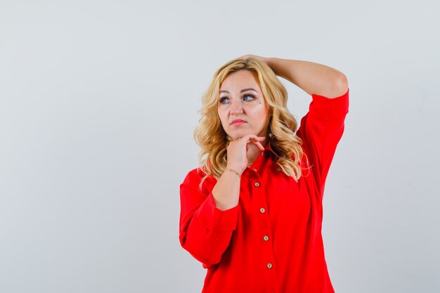 Blonde woman leaning chin on hand, putting hand on head and posing at camera in red blouse and looking pretty ,