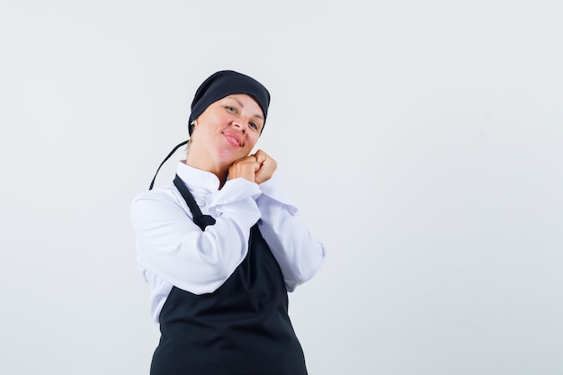 Blonde woman leaning chin on palms,clenching fists in black cook uniform and looking pretty.