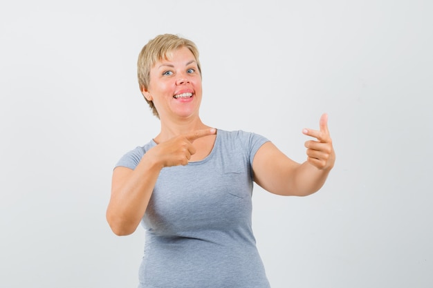 Blonde woman in light blue t-shirt pointing to the right corner and looking happy , front view.