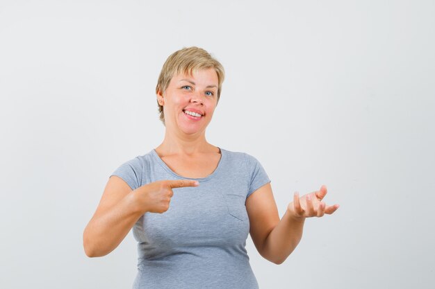 Blonde woman in light blue t-shirt pretending to hold something to hand pointing to it by other hand and looking cheerful , front view.