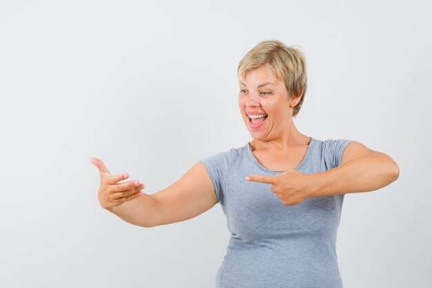 Blonde woman in light blue t-shirt pretending to look at texts on phone and pointing to her hand and looking cheerful , front view.