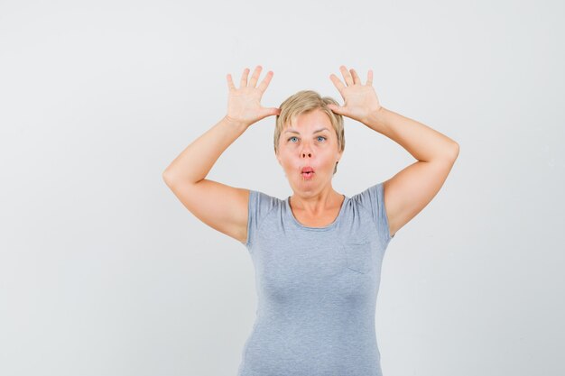 Blonde woman in light blue t-shirt raising her palms in surrender and looking surprised , front view.