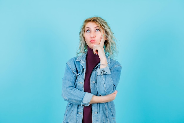 Blonde woman looking up by holding hand on cheek on blue background