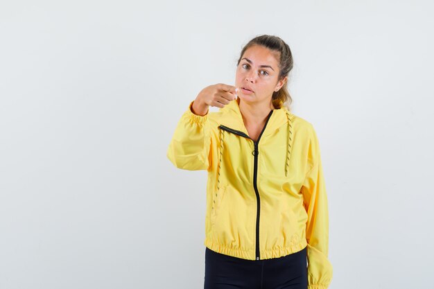 Blonde woman pointing at front with index finger in yellow bomber jacket and black pants and looking pretty