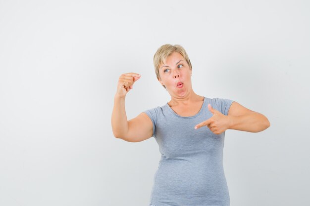 Blonde woman pointing to her muscles in light blue t-shirt and looking amazed , front view.